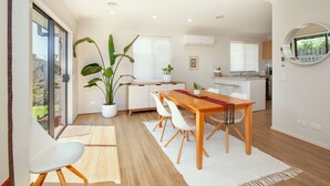 A modern kitchen with white cabinetry, a dining area with a wooden table and white chairs, and a large potted plant. - Northside Townhouse, Geelong (Norlane)