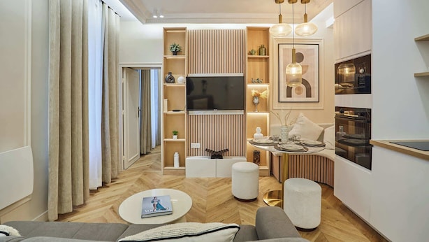 A modern living room with a flat-screen TV, wooden shelving, a dining area with a table set, and a kitchen with a stove.