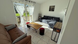 Un salon avec un canapé marron, une table basse en bois et un canapé gris. On y trouve une fenêtre avec des rideaux légers, une petite table avec des livres et un cadre photo noir accroché au mur.