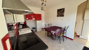 Une cuisine compacte équipée d’une cuisinière, d’un évier et de meubles de rangement rouges. Une table à manger avec des chaises est placée près d’une porte blanche.