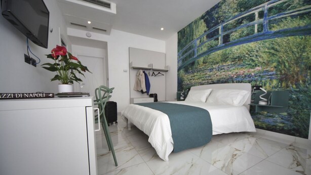 A modern hotel room with a bed, a desk with a plant, a TV, and a large painting on the wall. - Becoloured Water Lily Suite. Where it is possible to dream among the masterpieces of art. (Napoli)