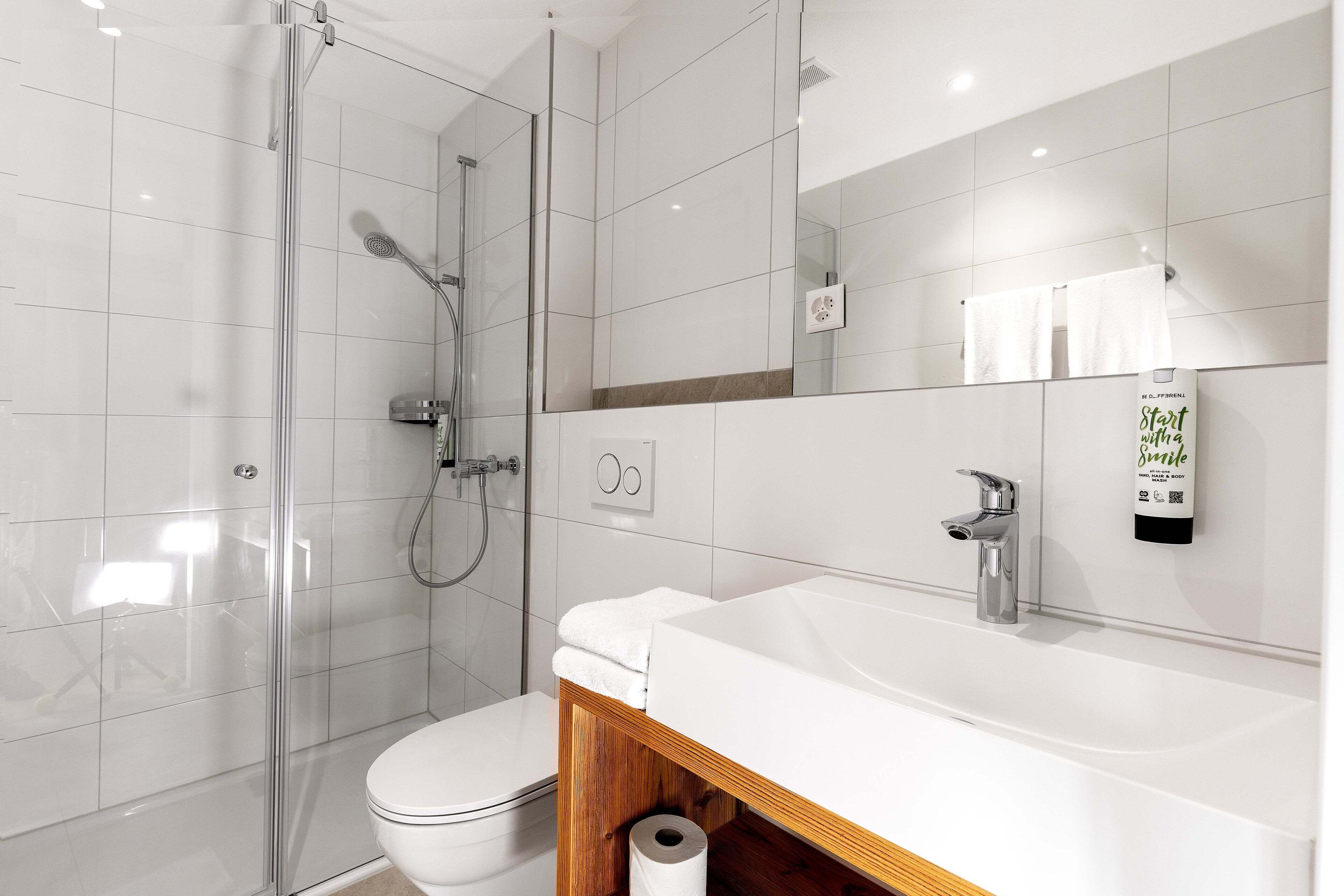 Un baño moderno con ducha envuelta en vidrio, inodoro blanco y un lavamanos amplio con espejo.
