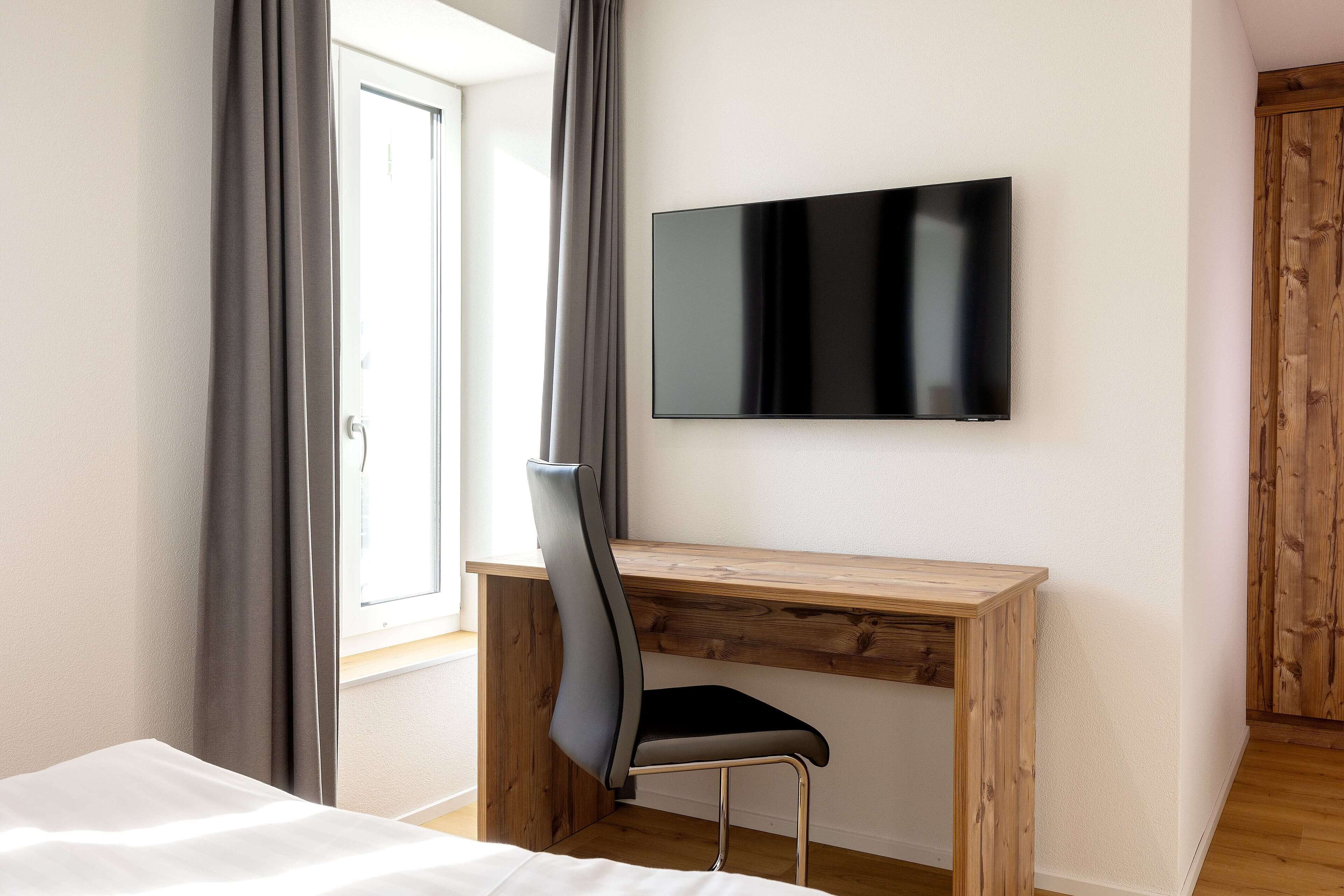 A bedroom with a bed, a desk with a chair, a TV mounted on the wall, and a window with curtains.