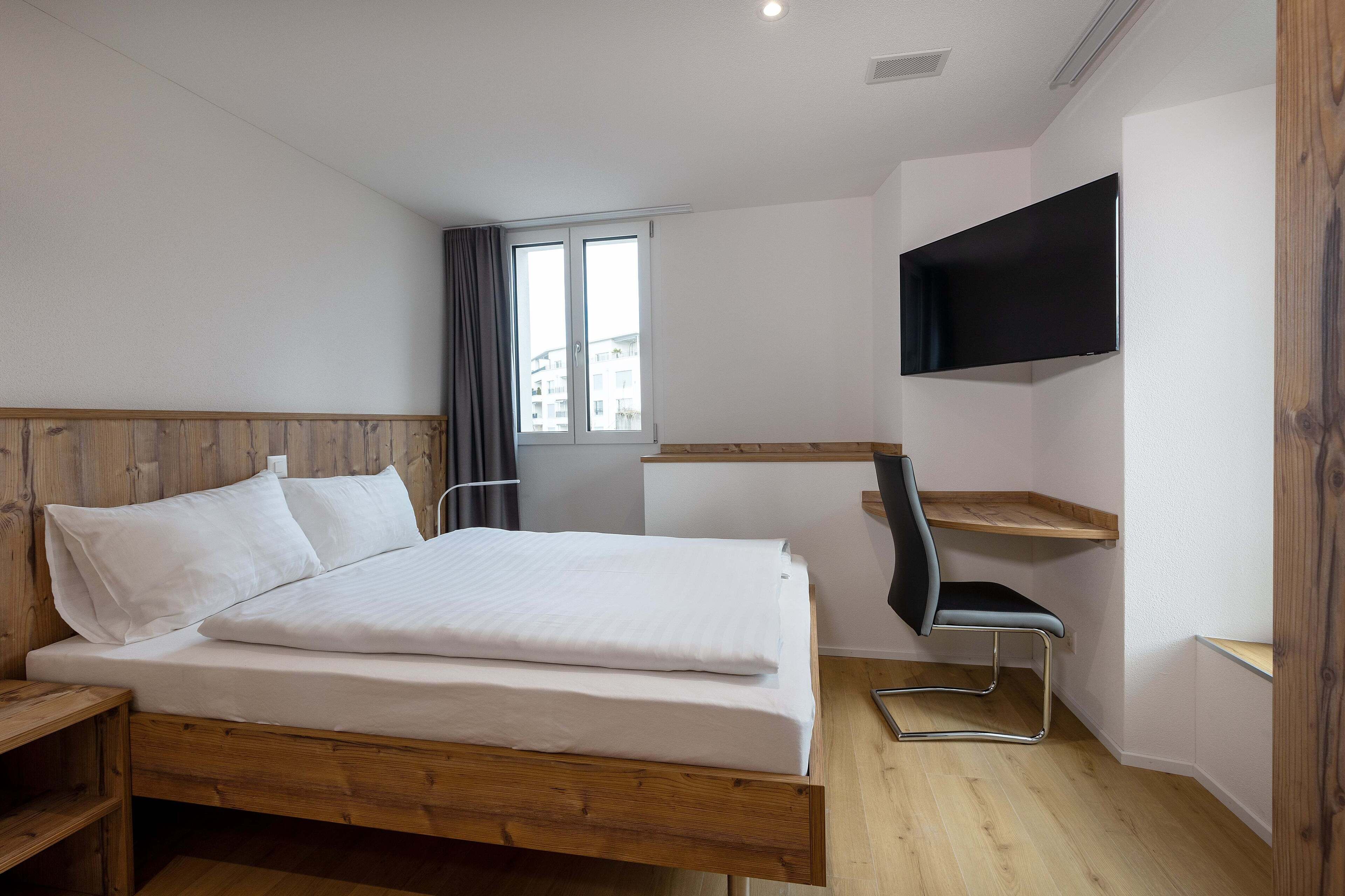 A modern hotel room with a wooden bed, a TV mounted on the wall, and a desk with a chair.