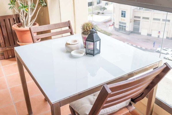 A balcony with a glass table, wooden chairs, a potted cactus, a lantern, and a view of the street.