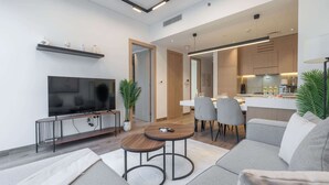 A modern living room with a flat-screen TV, a coffee table, and a sofa. There is a dining area with chairs and a kitchen in the background.