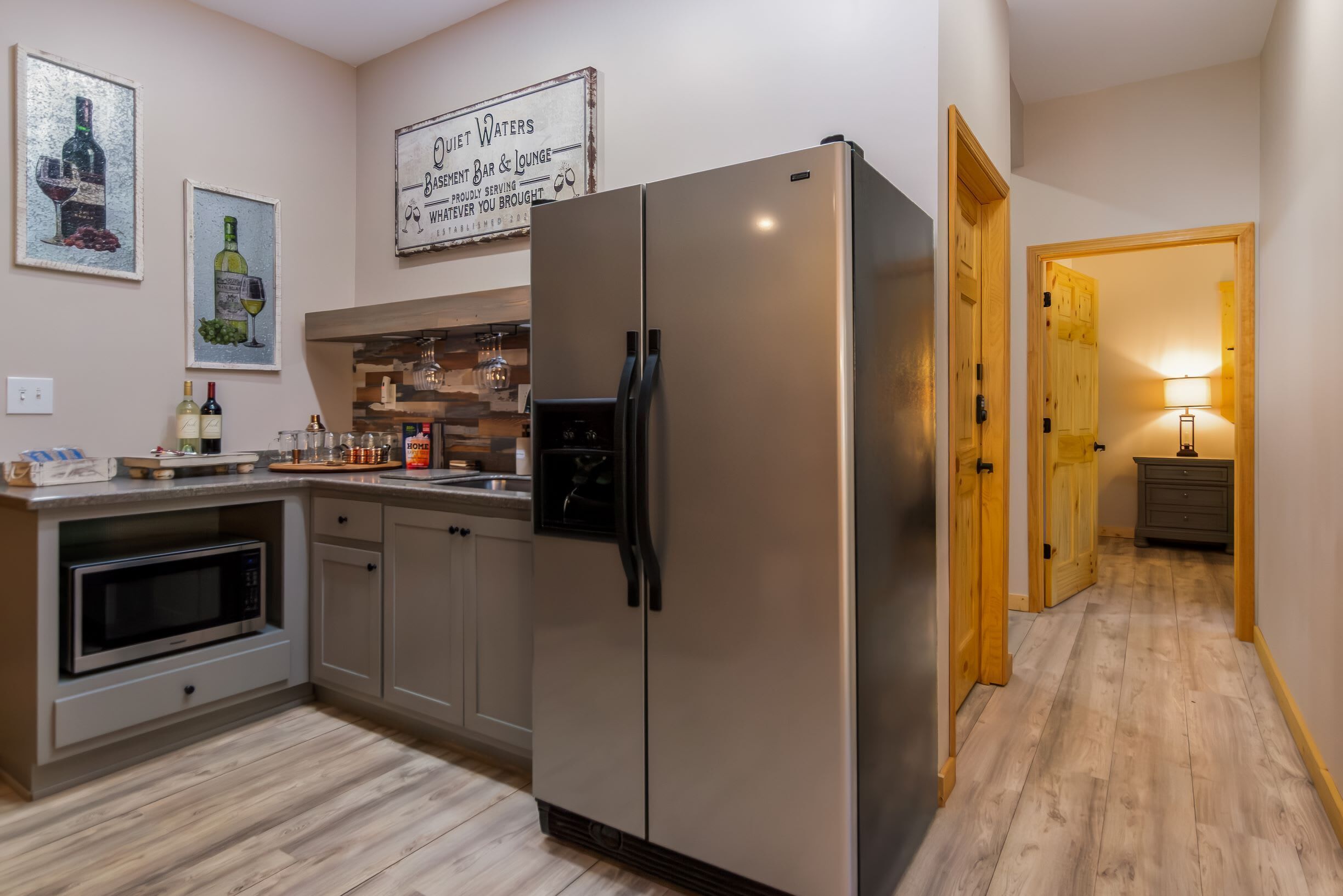 Una cocina moderna con un refrigerador de acero inoxidable, armarios de madera y un microondas. Hay botellas de vino y copas sobre la encimera, y un cartel sobre el refrigerador.