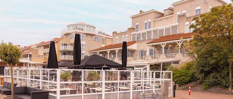 A waterfront restaurant with outdoor seating, a covered area, and a view of historic buildings.