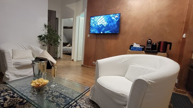 Una sala de estar con una mesa de centro de vidrio, sillones blancos, un televisor montado en la pared y piso de madera.