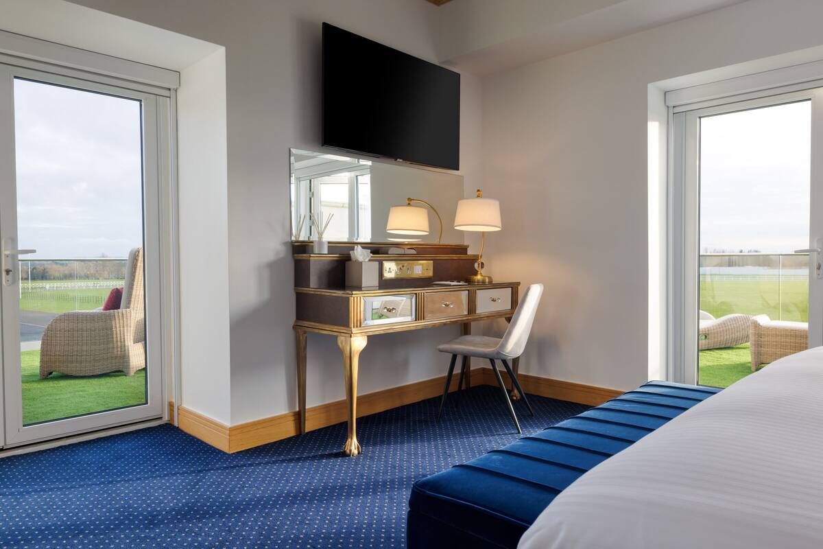 A hotel room with a large bed, a desk with a chair, a television mounted above the desk, and a view of the outdoors through the windows.