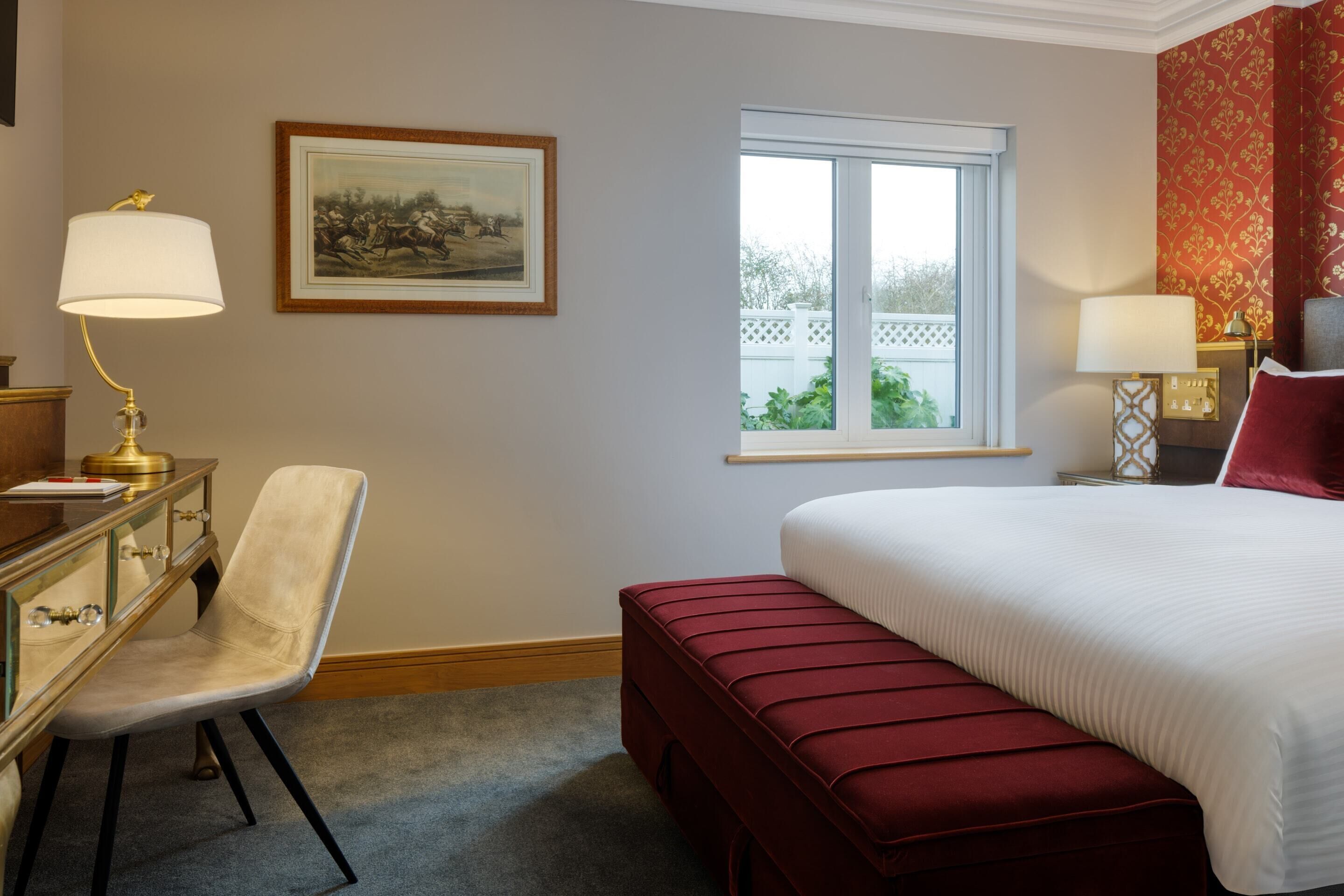 A hotel room with a bed, a desk with a lamp, a chair, a window with a view, and a framed picture on the wall.