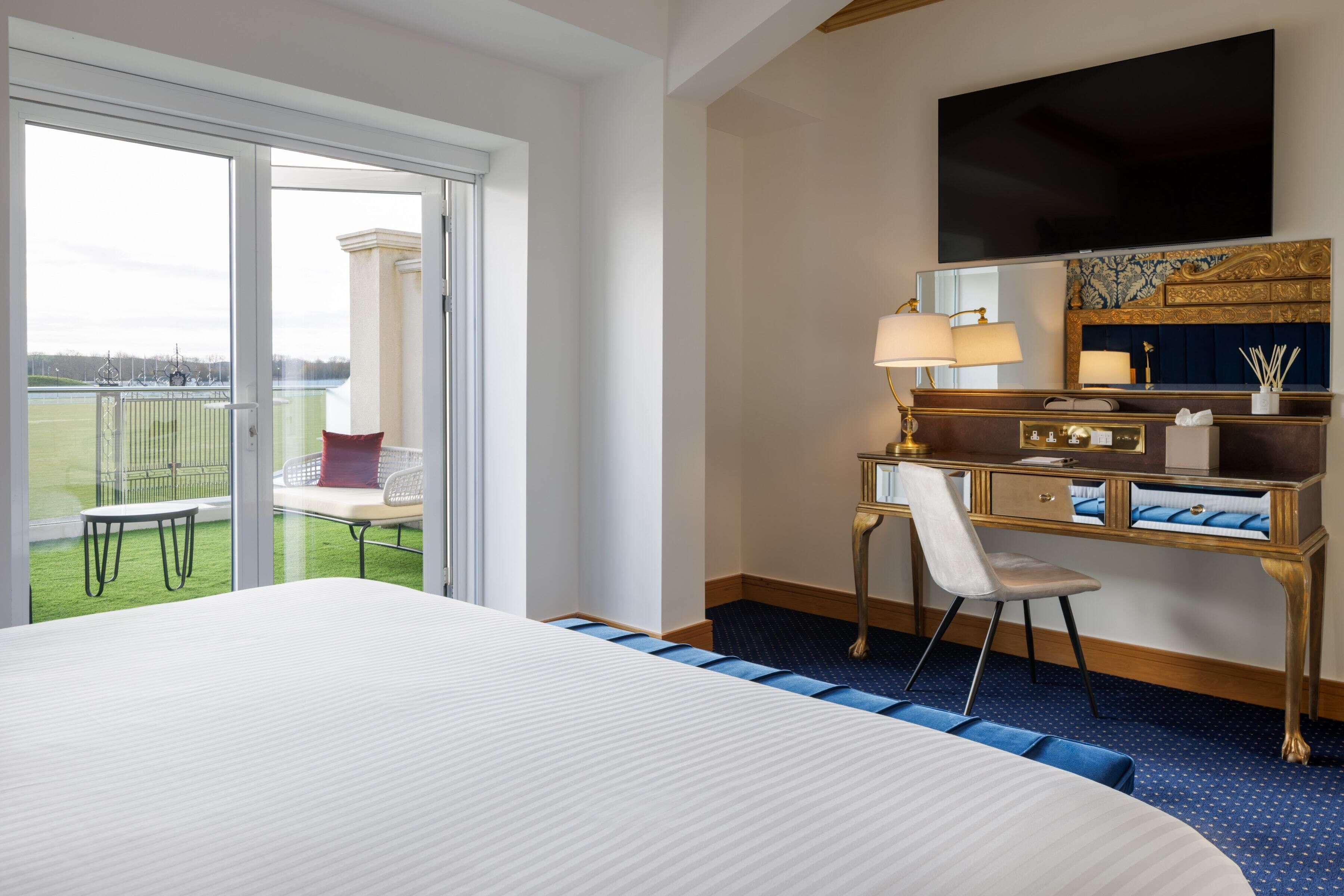 A hotel room with a large bed, a desk with a chair, a TV, and a balcony with a table and chairs.
