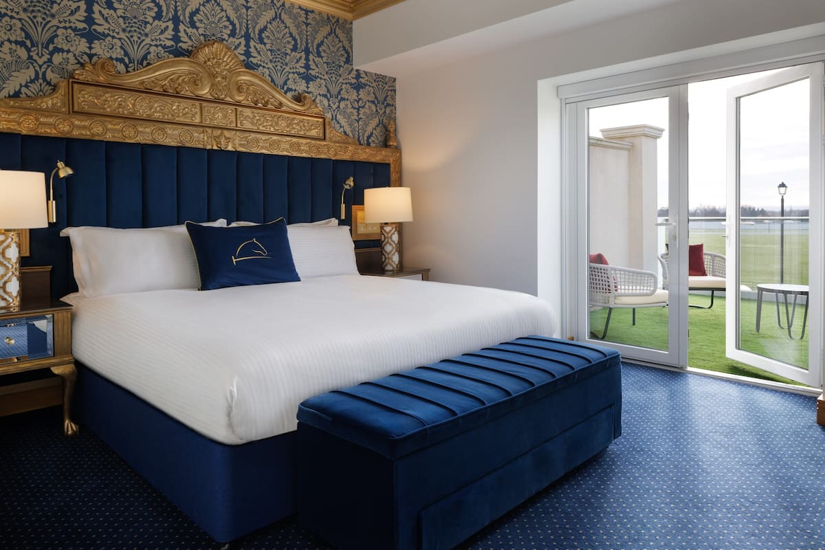 A hotel room with a large bed, a blue upholstered bench, a nightstand with a lamp, and a view of a tennis court through a glass door.