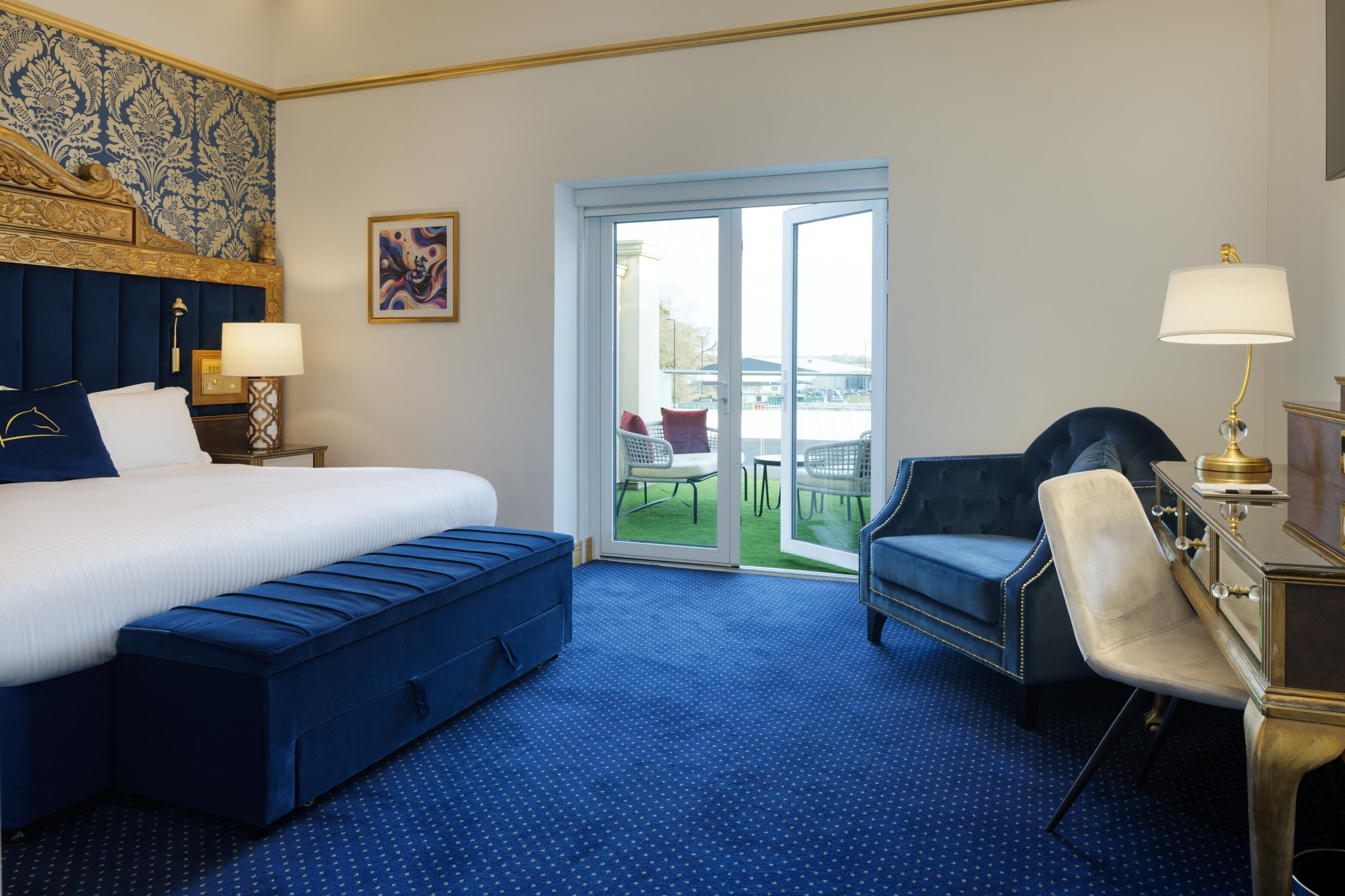 Una habitación de hotel con una cama grande, un banco azul, un escritorio con una lámpara y vistas al exterior a través de un ventanal.