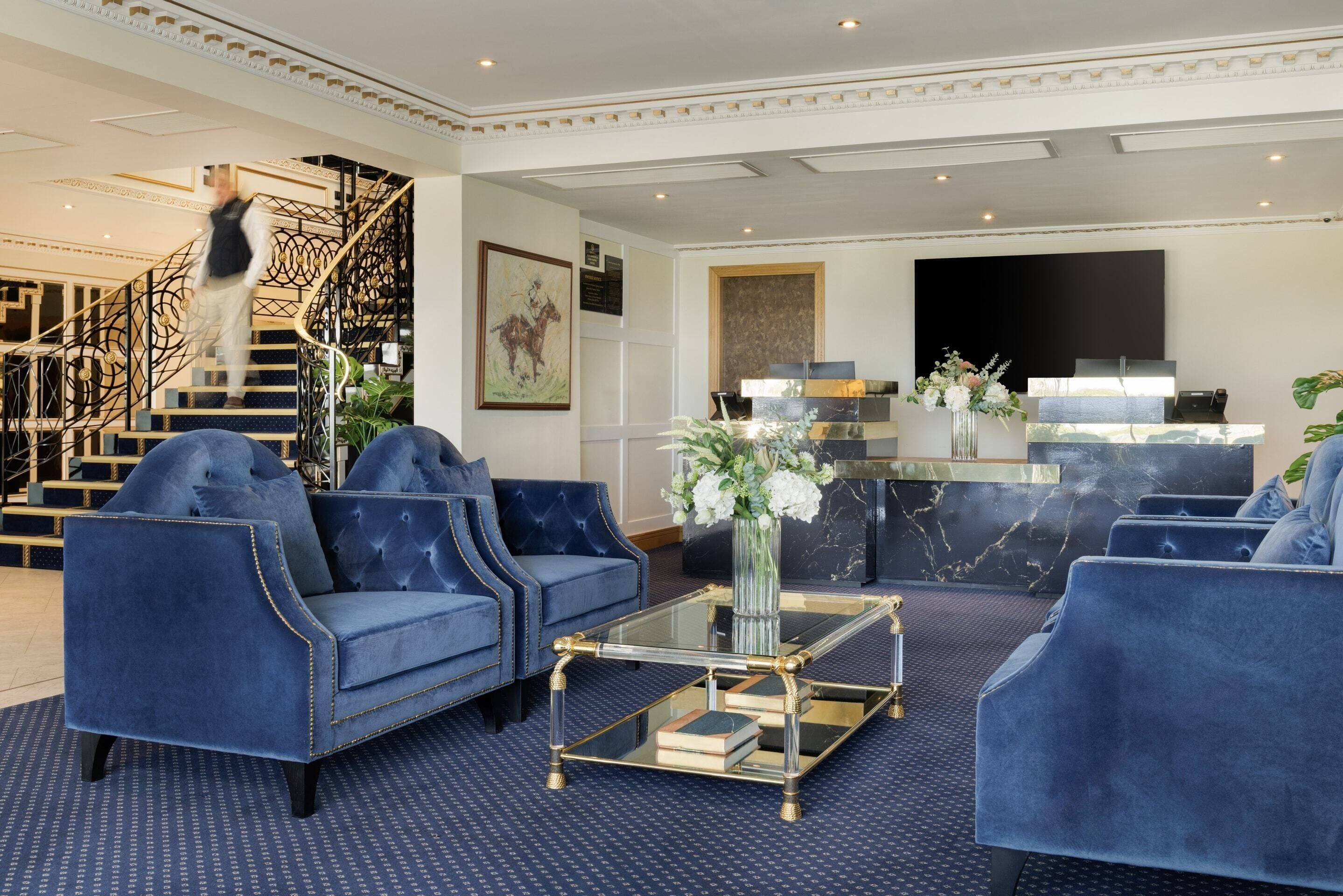 Een hal met blauwe gestoffeerde banken, een glazen salontafel, een receptiebalie en een trap.
