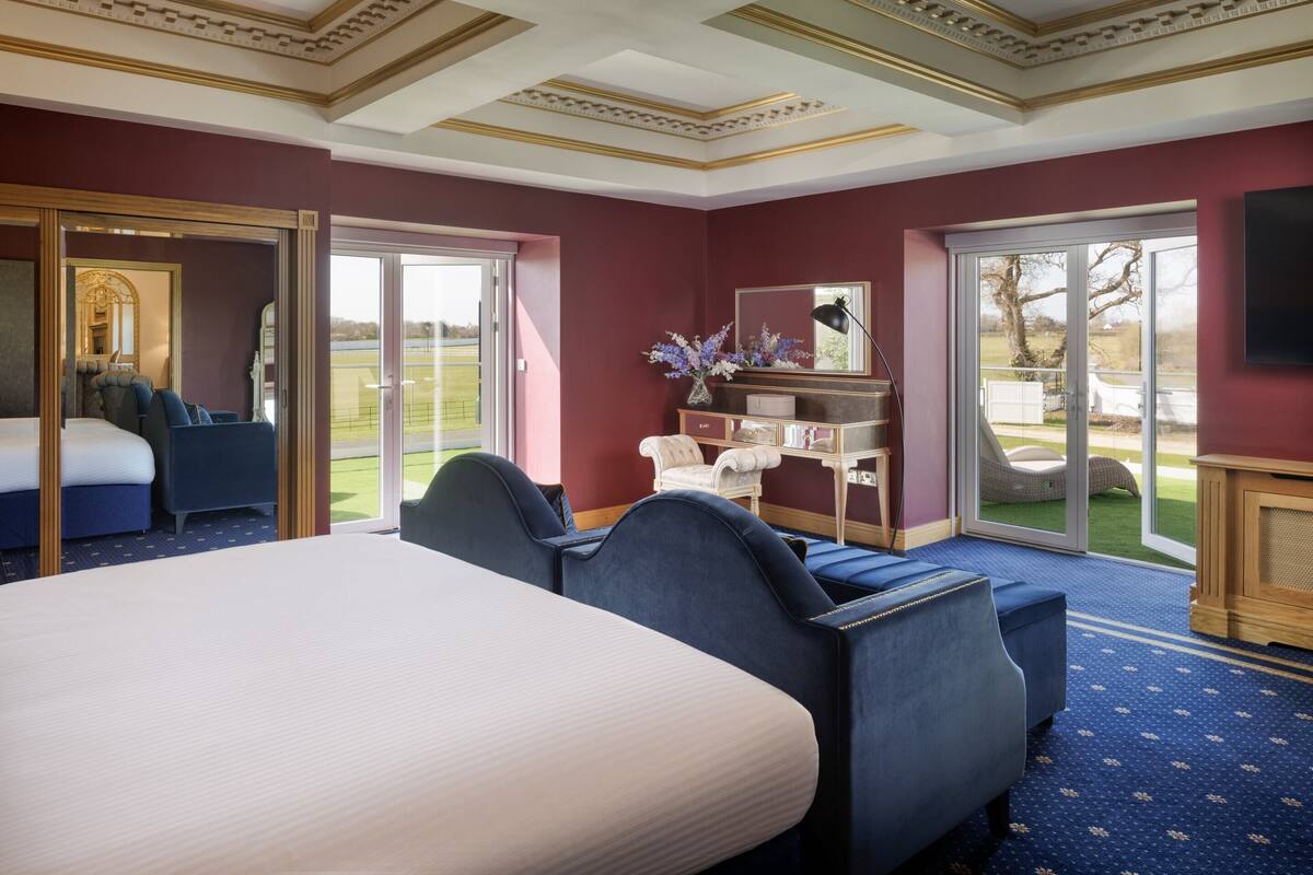 A hotel room with a large bed, a seating area with blue upholstered chairs, a small table, and a large window offering a view of the outdoors.