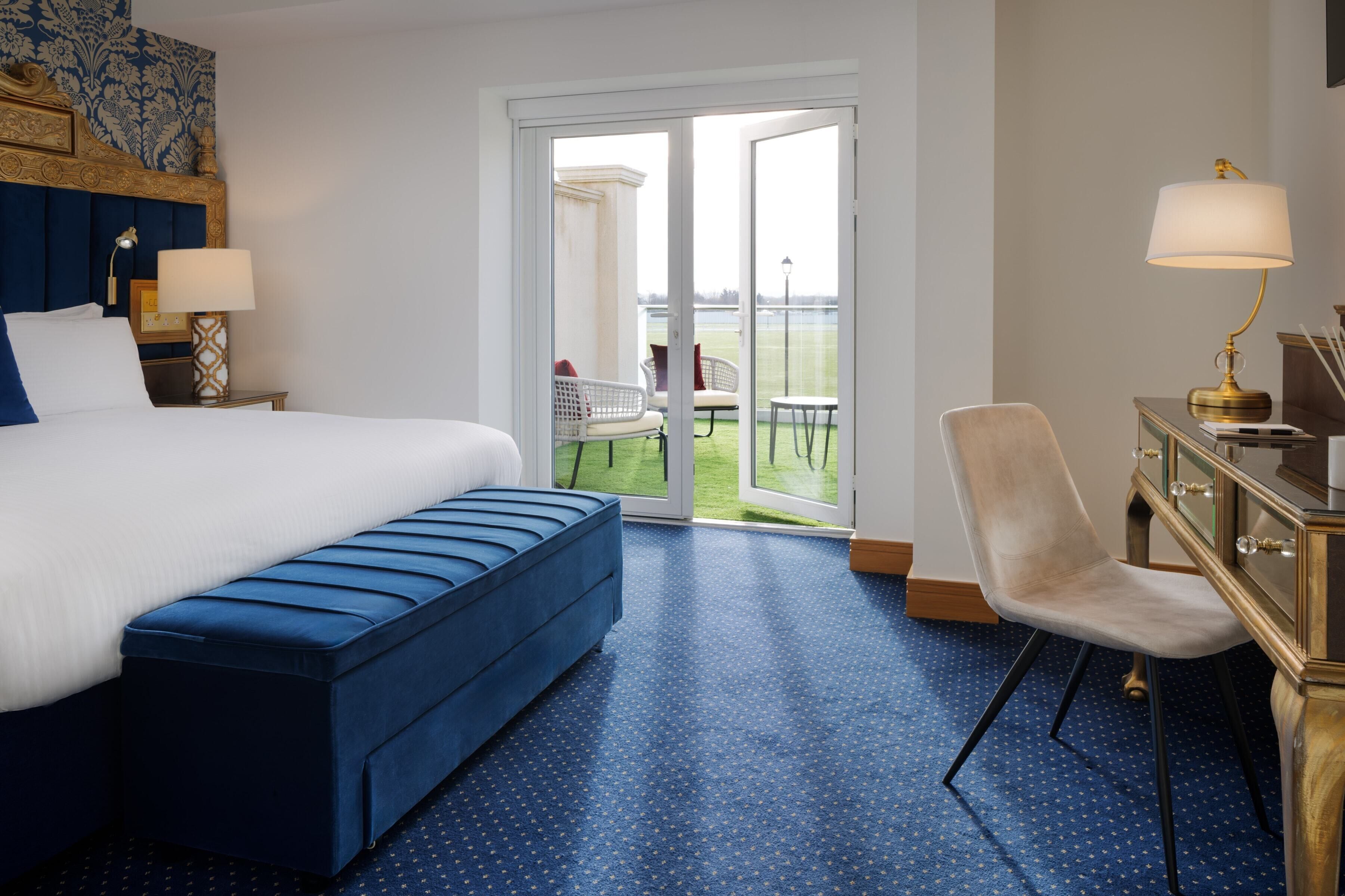 A hotel room with a large bed, a blue bench, a desk with a lamp, and a view of a tennis court through a glass door.