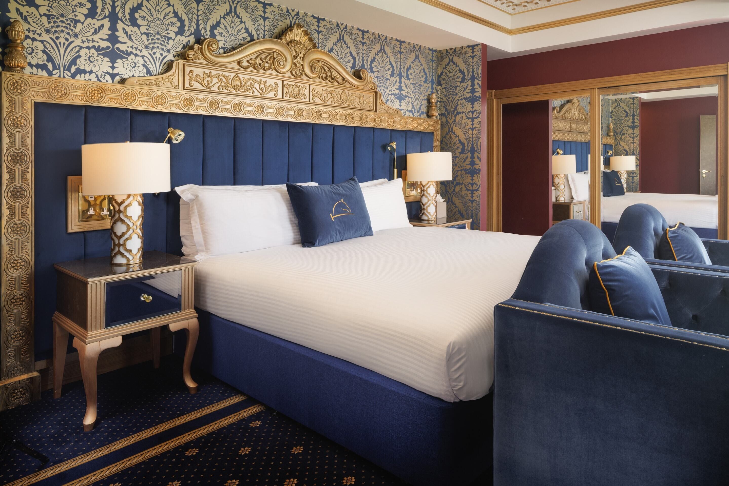 Quarto de hotel com uma cama grande, duas mesas de cabeceira com abajures, um espelho e uma poltrona azul estofada.