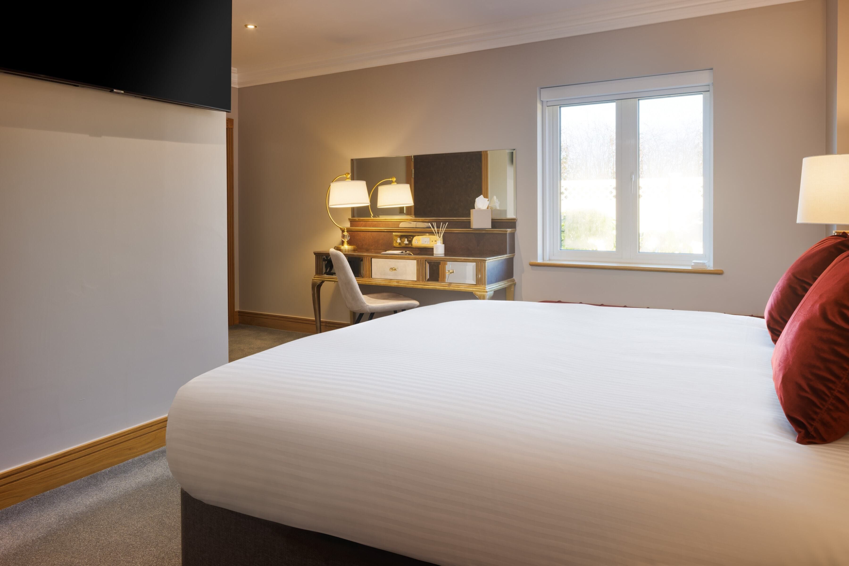 A hotel room with a large bed, a desk with a chair, a mirror, and a window.