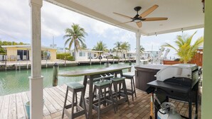Un espacio al aire libre techado con un bar, taburetes, una parrilla y vistas a un puerto deportivo con barcos y palmeras.