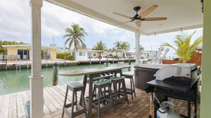 Un espace extérieur abrité comprenant un bar, des tabourets, un barbecue et une vue sur une marina avec des bateaux et des palmiers.