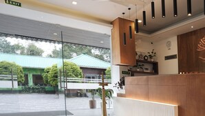 Un área de recepción moderna con un mostrador de madera, una lámpara de bronce y vistas a un edificio con techo verde a través de una puerta de cristal.