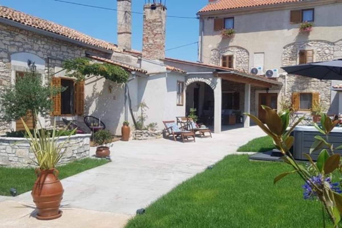 A two-story building with stone walls and a tiled roof. There is a covered patio area with outdoor furniture and a barbecue grill.