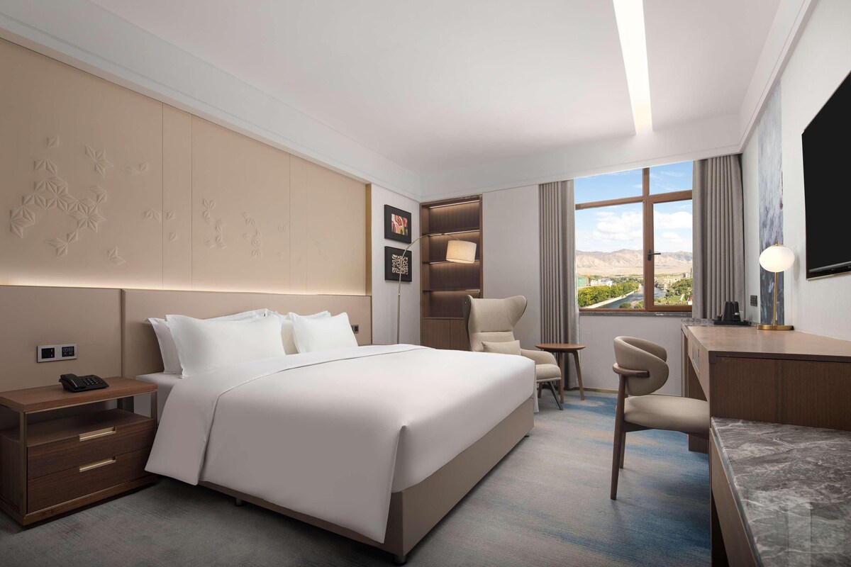 A modern hotel room with a large bed, a desk, a chair, a nightstand, and a TV.