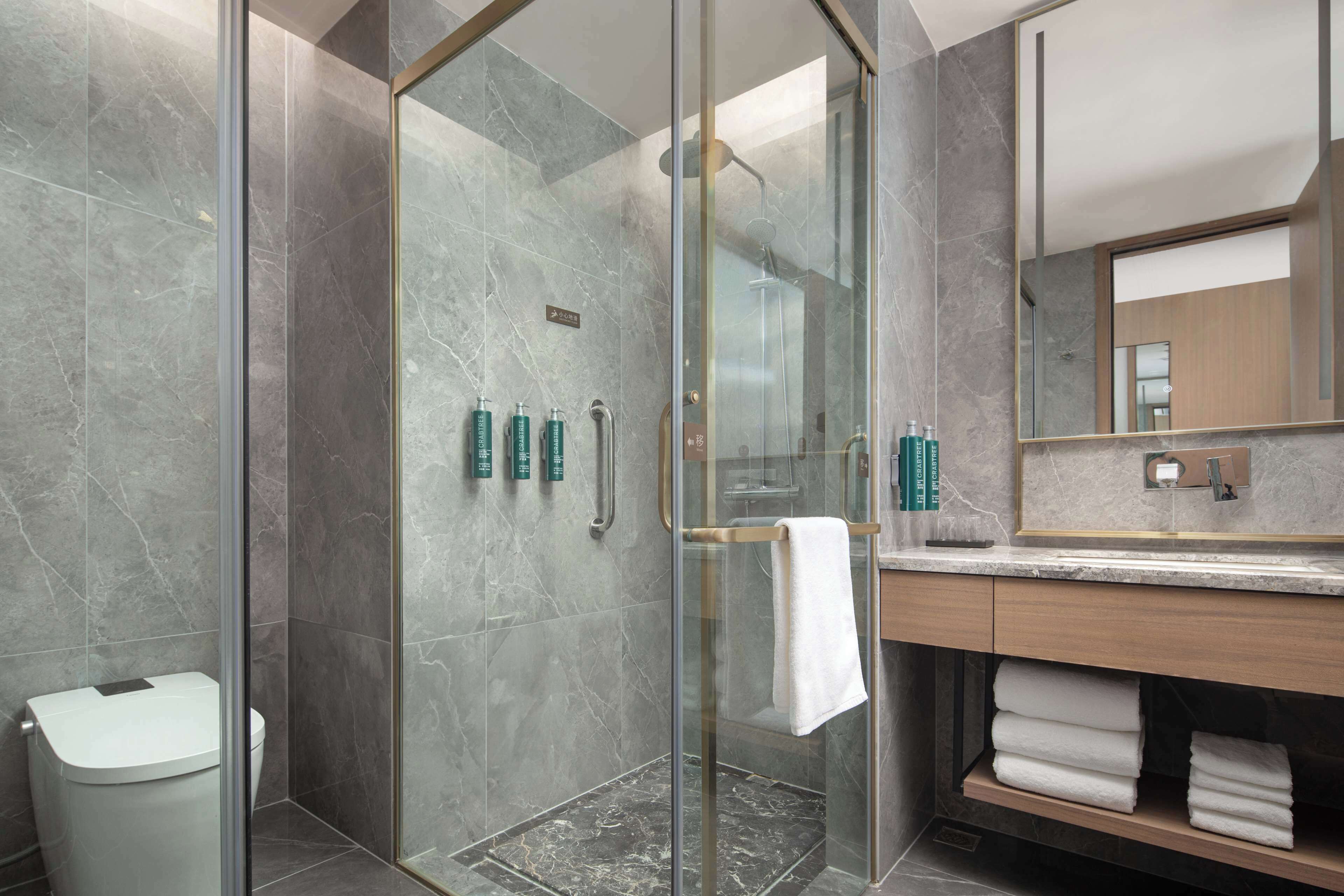 A modern bathroom with a glass-enclosed shower, marble tiles, a wooden vanity, and a large mirror.