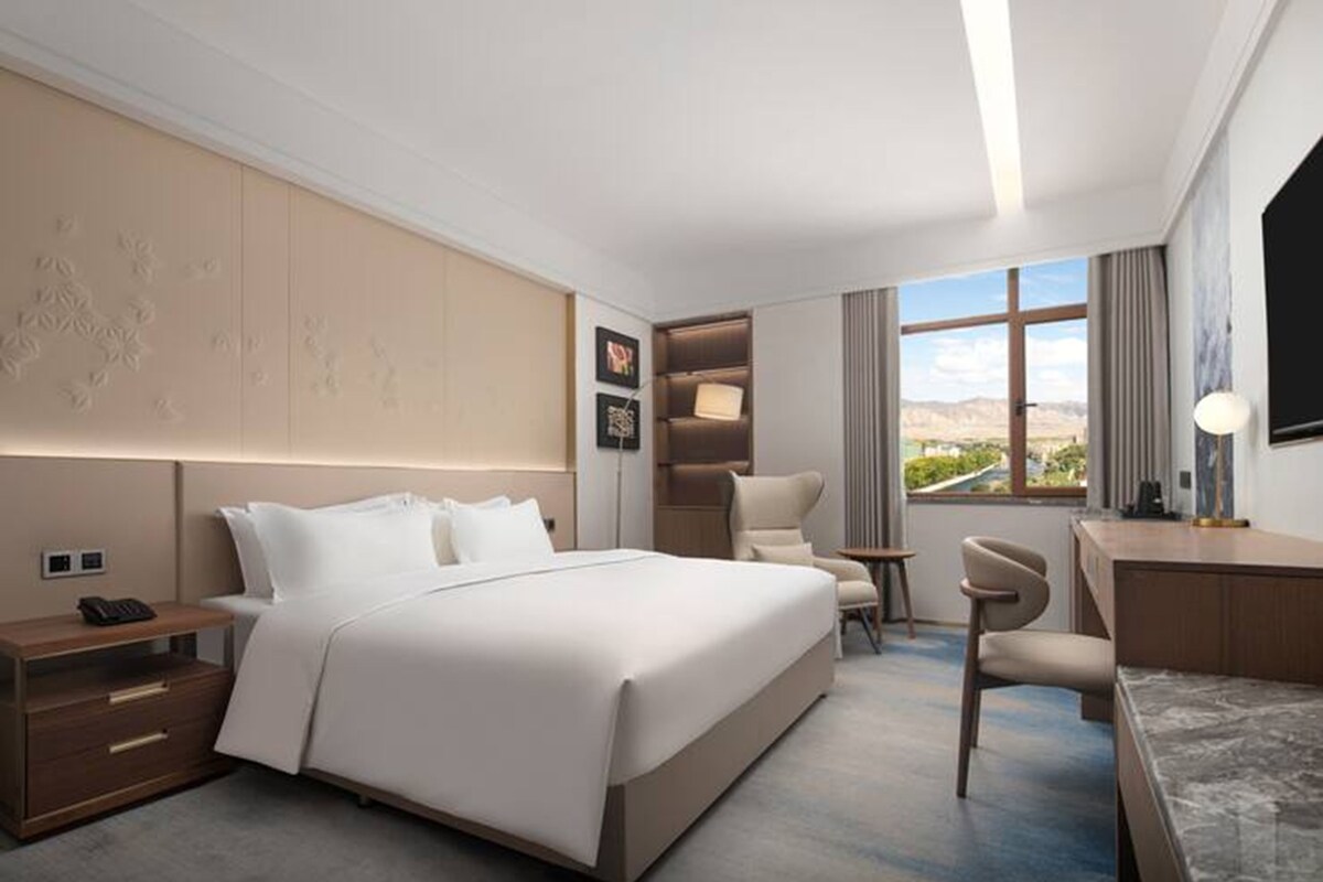 A modern hotel room with a large bed, a desk, a chair, a small table, and a view of the outdoors.