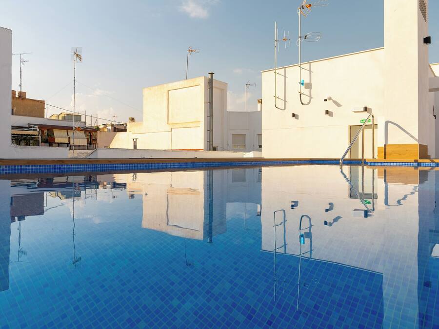 Une piscine d’un bleu éclatant, bordée de carreaux, entourée de murs blancs et de balcons ornés de diverses antennes et de caméras de surveillance.