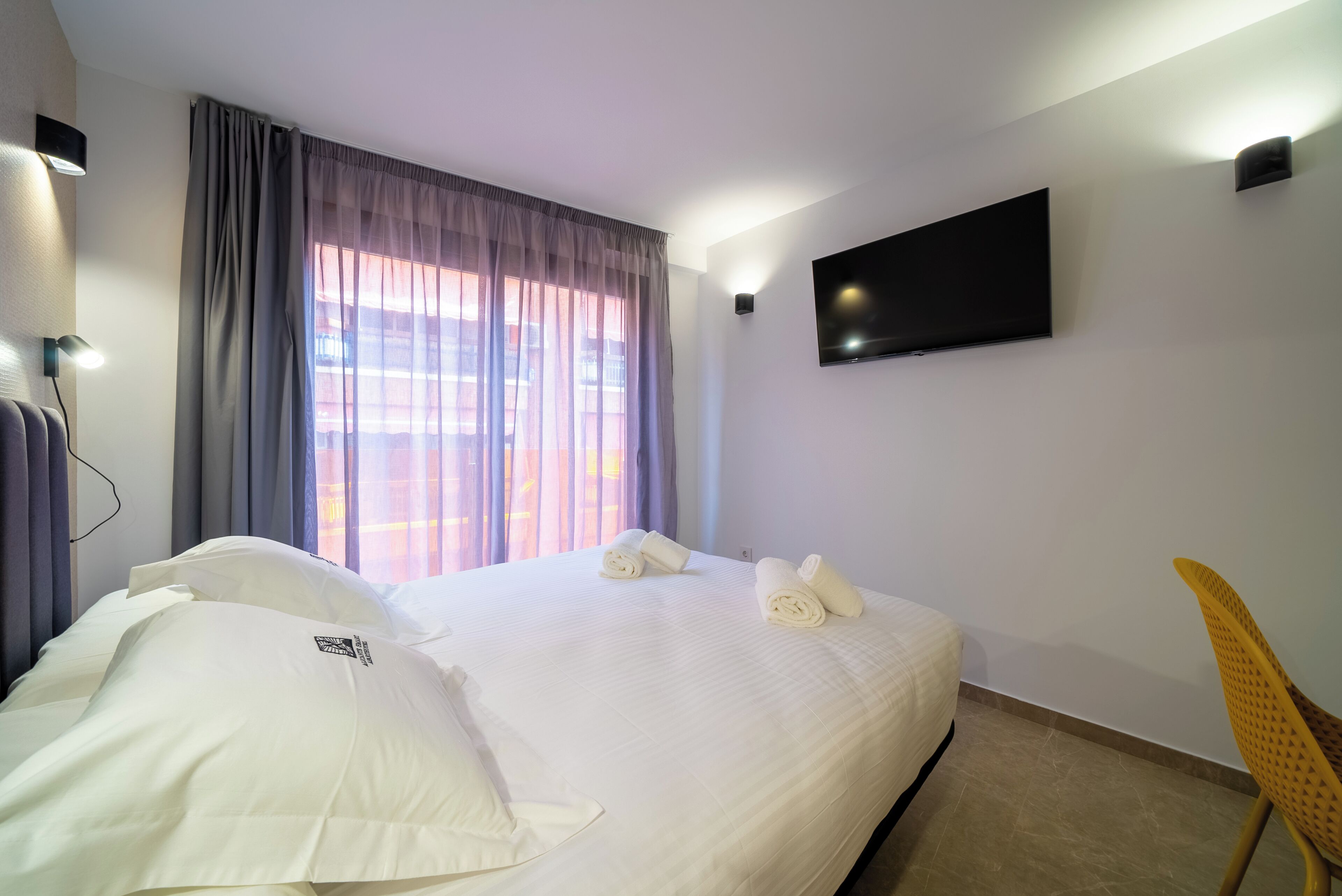 Una habitación de hotel con una cama, un televisor montado en la pared y una silla.
