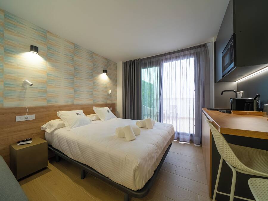 Una habitación de hotel moderna con una cama bien hecha, una mesita de noche pequeña, un escritorio con una silla y un ventanal con cortinas.