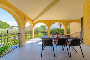 Un portico coperto con tavolo e sedie, un parapetto e vista su aree verdi ed edifici.