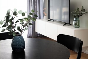 Moderni olohuone, jossa on tumma puinen pöytä, sininen maljakko, jossa on vihreitä lehtiä, musta tuoli, litteänäyttöinen televisio ja valkoinen kaappi.