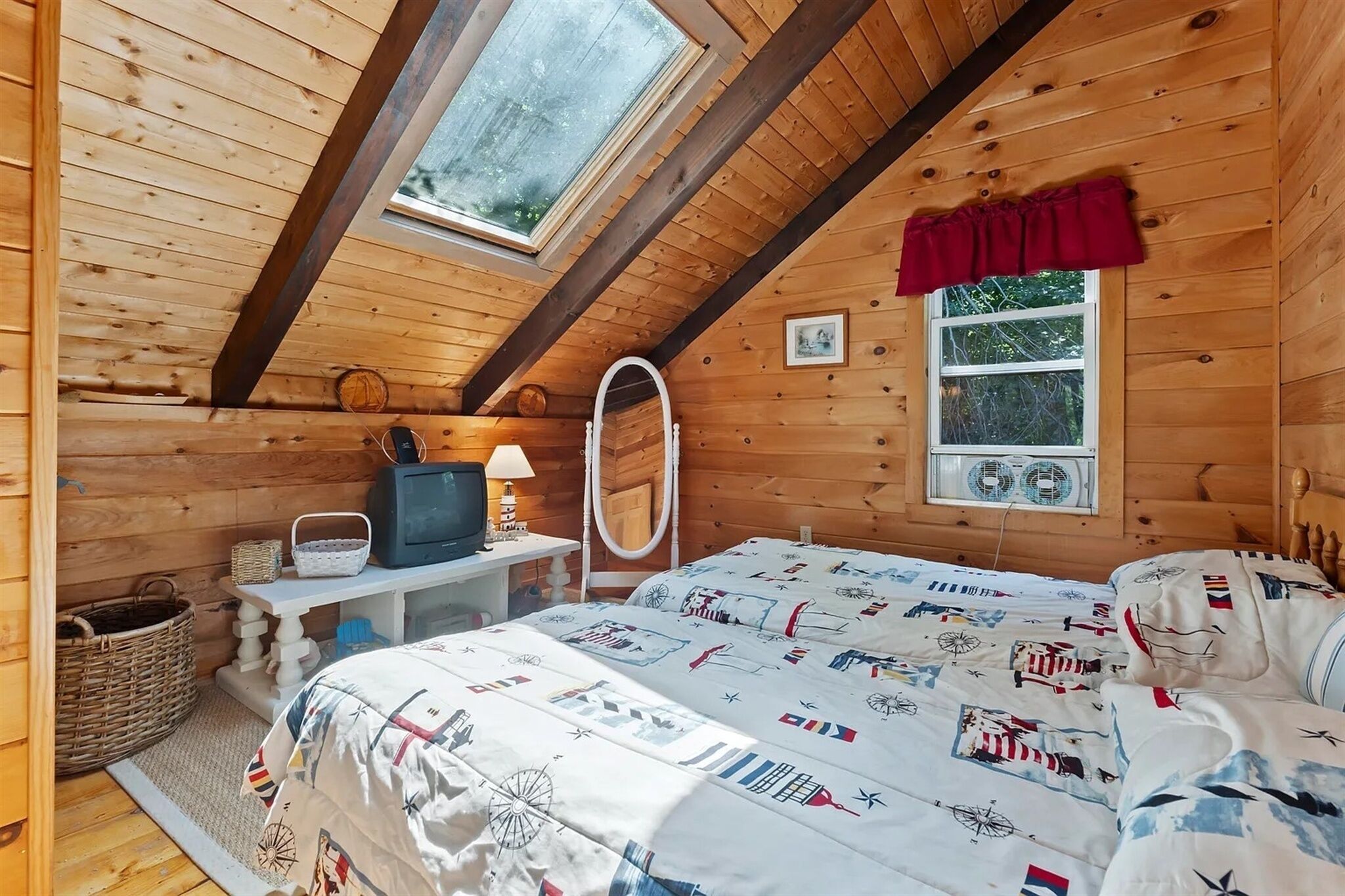 A cozy wooden cabin bedroom with a sloped ceiling, a bed with a nautical-themed comforter, a television, and a small table with a lamp.