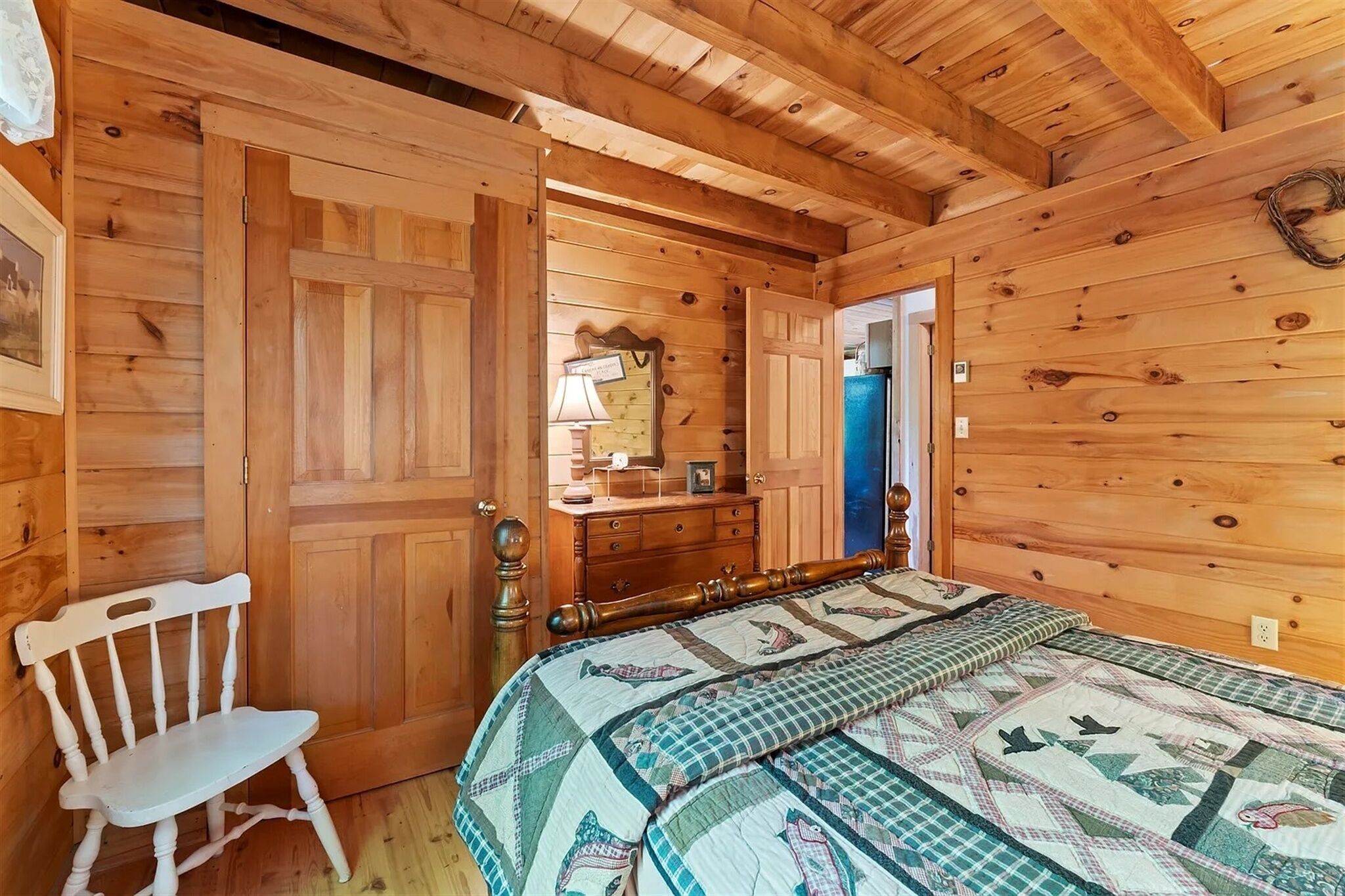 Een slaapkamer in een houten blokhut met een bed, een kaptafel, een stoel en een deur naar een andere kamer.