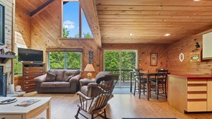 A cozy cabin interior with a wooden ceiling, a television, a sofa, a rocking chair, and a dining area with a table and chairs. - Unusual Latitude Limit 7 3 Bedroom Villa (Raymond)