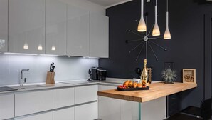 A modern kitchen with white cabinets, a wooden countertop, and a black backsplash. There are hanging pendant lights, a coffee machine, and a bottle of champagne on the counter. - Villa Luxor With Heated Pool (Malinska-Dubasnica)