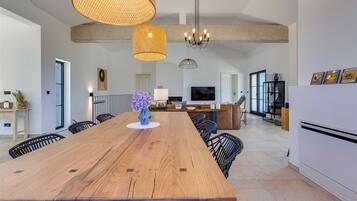 A spacious dining area with a long wooden table, black chairs, and a vase of purple flowers. There are hanging wicker lamps and a chandelier.