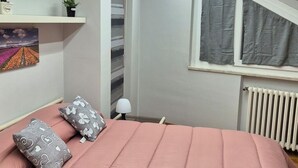 Makuuhuone, jossa on vaaleanpunainen sänky, sydänkuvioilla koristellut tyynyt, pieni pöytä lampun kanssa, patteri ja harmailla verhoilla varustettu ikkuna.