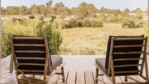Two wooden lounge chairs with cushions on a wooden deck overlooking a grassy field with trees and a clear sky. - Casas de Arroz - Cabin (Possanco - Comporta)
