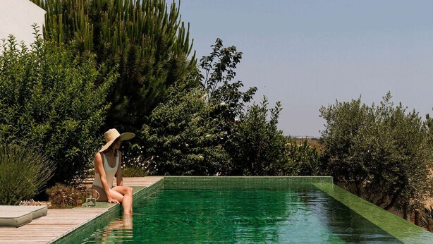 A person sitting by a clear, green pool surrounded by lush greenery.