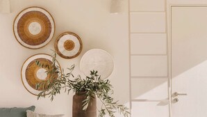 A white table with a plant in a brown vase, a wooden cutting board with apples, and a white wall with three woven wall hangings.