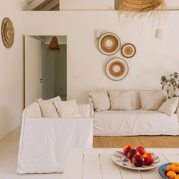 A minimalist living room with a white sofa, a woven wall hanging, and a fruit bowl on the table.