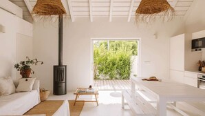 A modern, minimalist living space with a white color scheme, featuring a sofa, a table, a bench, a fireplace, and a view of greenery outside.