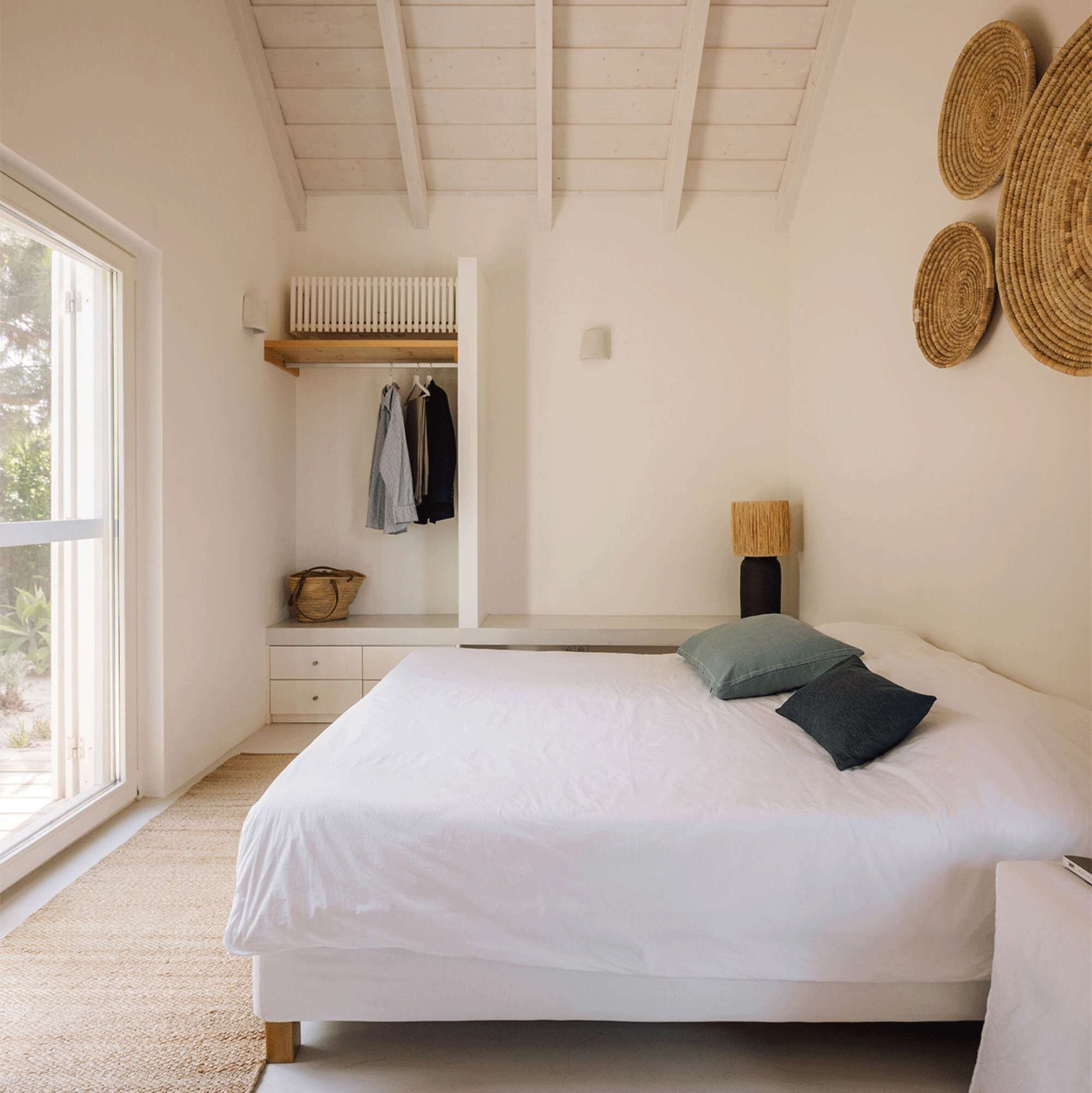 Quarto com cama, mesa de cabeceira, abajur, luminária pendente, janela e armário com roupas.