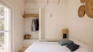 A bedroom with a bed, a bedside table, a lamp, a hanging light fixture, a window, and a closet with clothes.