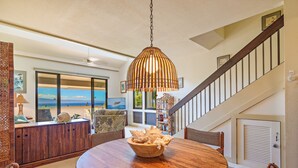 A dining area with a wooden table and chairs, a hanging light fixture, and a view of the ocean through large windows.