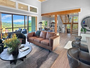 A spacious living area with a large window offering a view of the outdoors, a brown leather sofa, a round coffee table, and a staircase leading to an upper level. - Mountain Modern 3BR cabin with hot tub, fire pit, lake access free train and rafting (Bryson City)